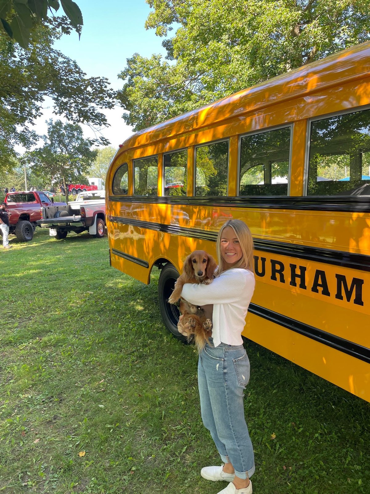 Woman holding a dog in front of a yellow school bus with Durham text on its side in a park setting.